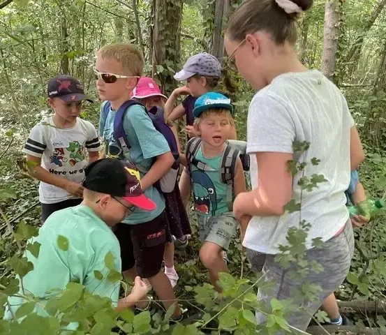 Eine Gruppe von Kindern, alle mit Mützen und Brillen, erkunden einen Wald. Ein Kind hockt sich hin und untersucht etwas, während andere zuschauen.