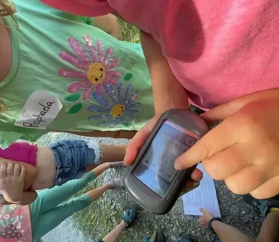 Mehrere Kinder in bunter Kleidung sind um eine Person versammelt, die ein Gerät, möglicherweise ein GPS, hält. Sie sind an einer Outdoor-Aktivität beteiligt.