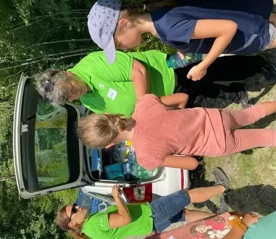 Eine Gruppe von Menschen in grünen und rosa Shirts lädt Gegenstände in den Kofferraum eines Autos in einem Waldgebiet.