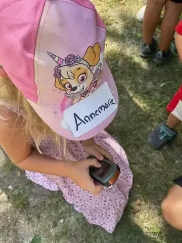 Ein Mädchen namens Annemarie sitzt auf dem Gras, hält ein kleines Objekt in der Hand und trägt einen rosa Hut mit einem Cartoon-Hundecharakter.