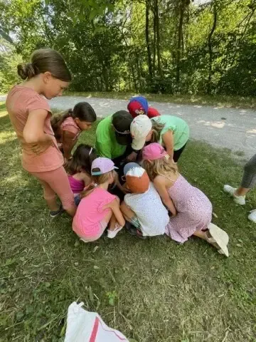 Mehrere junge Mädchen und zwei Erwachsene hockten sich auf einem Grasfeld und sahen sich etwas auf dem Boden an. Sie sind in Freizeitkleidung gekleidet und scheinen an einer Outdoor-Aktivität beteiligt zu sein.