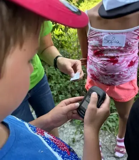 Ein Kind hält ein Gerät, während ein anderes Kind ein Papier und eine Karte in der Hand hat, mit einem Mädchen namens Sofia im Hintergrund.