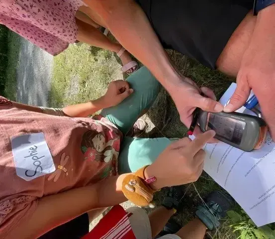 Mehrere Menschen sitzen auf dem Gras mit einem Papier und einem Telefon. Ein Mädchen namens Sophie hält ein Telefon.