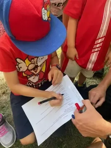 Ein junges Kind in einem roten Shirt und Hut unterschreibt ein Blatt Papier im Freien.