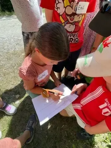 Ein Mädchen mit Brille unterschreibt ein Papier, während es auf dem Gras mit einem Jungen in einem roten T-Shirt mit weißen Streifen sitzt.