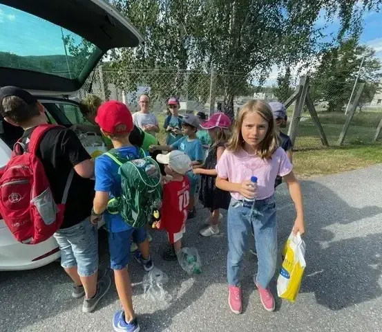 Eine Gruppe von Kindern und Erwachsenen steht neben einem Auto mit offenem Kofferraum in einem Außenbereich mit Bäumen und einem Zaun.