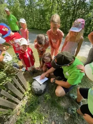 Eine Gruppe von Kindern und Erwachsenen ist um einen Felsen versammelt und nimmt wahrscheinlich an einer Outdoor-Bildungsaktivität teil. Ein Mann in Grün kniet und hält ein Papier, während Kinder und Erwachsene beobachten. Bäume sind im Hintergrund.