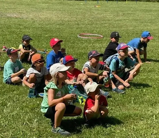 Kinder in Mützen und Turnschuhen sitzen auf einem Grasfeld, einige halten Wasserflaschen. Ein roter Hula-Hoop liegt in der Nähe. Die Schatten der Kinder werden auf das Gras geworfen.