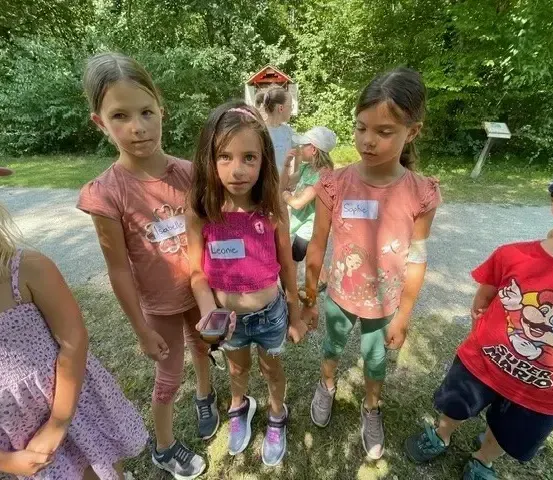 Mehrere Kinder stehen auf Gras, eines hält ein Telefon. Eine Mädchen namens Leonie zeigt ihren Bauch. Das Mädchen neben ihr heißt Sophie. Ein kleines Mädchen mit Hut steht etwas hinter ihnen.