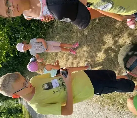 Eine Gruppe von Menschen, einschließlich Kindern und Erwachsenen, steht auf einem schotterigen Pfad mit Gras und Pflanzen an der Seite. Sie tragen verschiedenfarbige Hemden und Mützen. Der Junge vorne hält ein Handy.