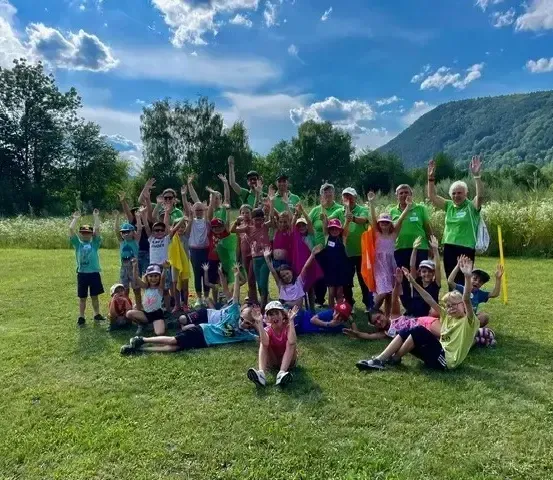 Eine große Gruppe von Menschen, einschließlich Erwachsener und Kindern, posiert für ein Foto auf einer Wiese mit einer Kulisse aus Bäumen und Bergen.