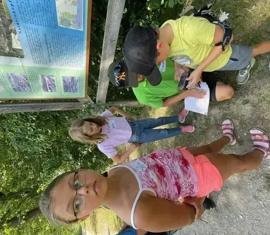 Vier Kinder sitzen im Freien neben einem Schild mit Bildungsinhalten. Sie sitzen auf dem Boden, schauen auf ein Papier und schreiben möglicherweise.