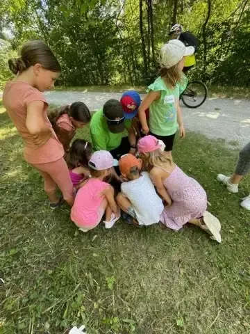 Eine Gruppe von Kindern ist auf dem Gras versammelt und schaut mit Erwachsenen etwas an. Ein Erwachsener trägt ein grünes Shirt. Ein Fahrrad ist im Hintergrund.