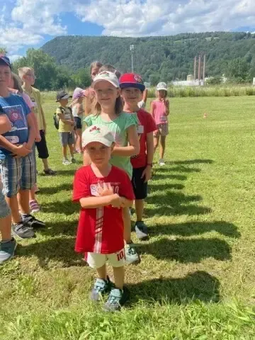 Eine Gruppe von Kindern steht auf einem Rasenfeld, einige tragen Mützen und Turnschuhe. Das Kind im roten T-Shirt schaut in die Kamera.
