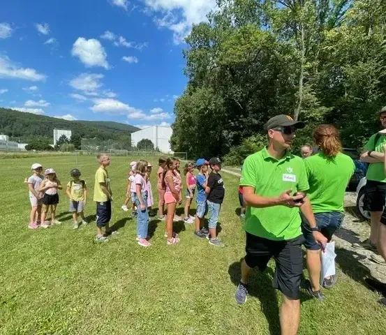 Eine Gruppe von Kindern steht in einem Grasfeld unter einem blauen Himmel. Sie tragen Mützen und Turnschuhe. Ein Mann in einem grünen Polohemd steht vor ihnen. Im Hintergrund befinden sich Bäume und ein Gebäude.
