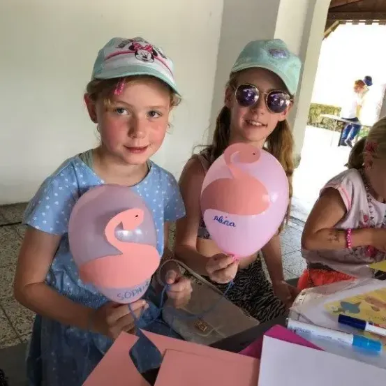 Zwei Mädchen halten rosa Flamingo-förmige Luftballons. Sie tragen Mützen und Sonnenbrillen. Andere Kinder sind im Hintergrund. Ein Tisch mit Bastelarbeiten und Papieren ist im Vordergrund.