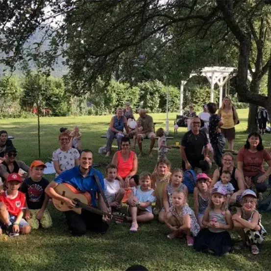 Eine Gruppe von Menschen, einschließlich Kindern, versammelt sich in einem Park. Einige sitzen auf dem Gras, während andere stehen. Ein Mann spielt Gitarre. Im Hintergrund gibt es Bäume, ein Pavillon und Berge.