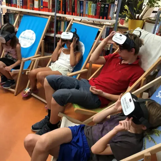 Vier Kinder in einer Bibliothek mit VR-Headsets sitzen in Liegestühlen und erkunden eine virtuelle Welt.