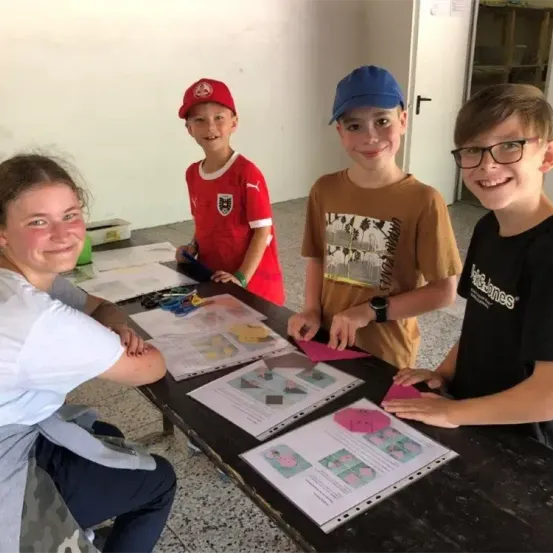 Drei Jungen und eine Frau sitzen an einem Tisch. Die Jungen lächeln und schauen in die Kamera. Die Frau lächelt und schaut auf den Tisch. Papiere, Scheren und ein Buch liegen auf dem Tisch.