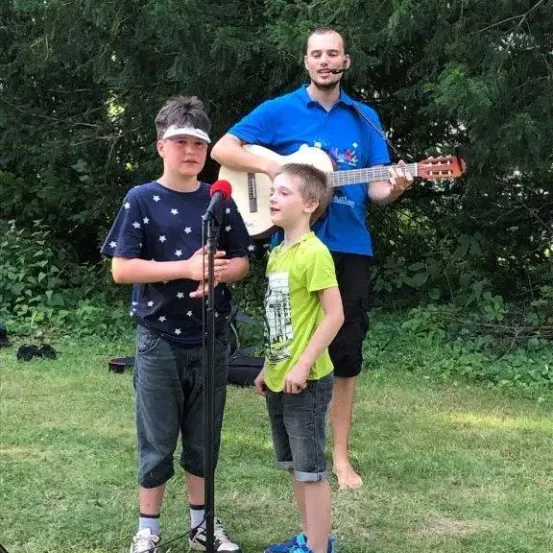 Zwei Jungen singen in ein Mikrofon, während ein Mann Gitarre spielt. Sie sind draußen in einem Grasbereich.