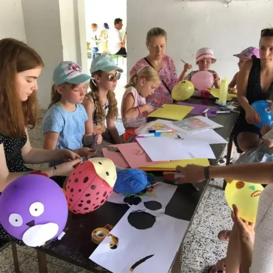 Mehrere Kinder und Erwachsene versammeln sich um einen langen Tisch. Sie beschäftigen sich mit Bastelarbeiten, mit Luftballons, Garn und Papieren auf dem Tisch. Die Kinder tragen Hüte und Sonnenbrillen, während auch Erwachsene anwesend sind.