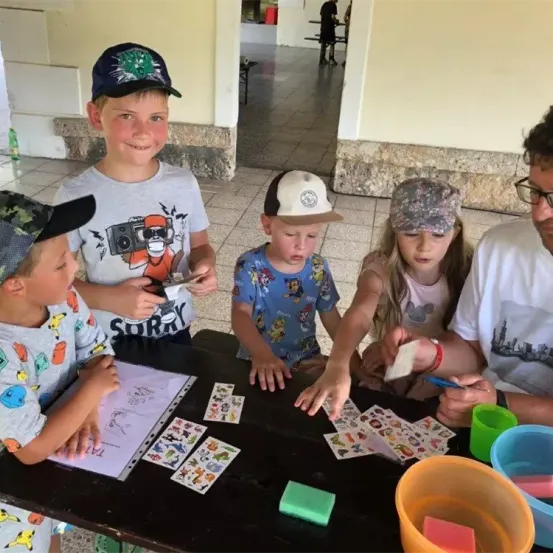 Vier Kinder und ein Erwachsener sitzen um einen Tisch herum und beschäftigen sich mit einer Bastelaktivität. Die Kinder tragen Mützen und T-Shirts mit verschiedenen Aufdrucken. Sie haben einen Tisch voller Bastelmaterial und Zubehör.