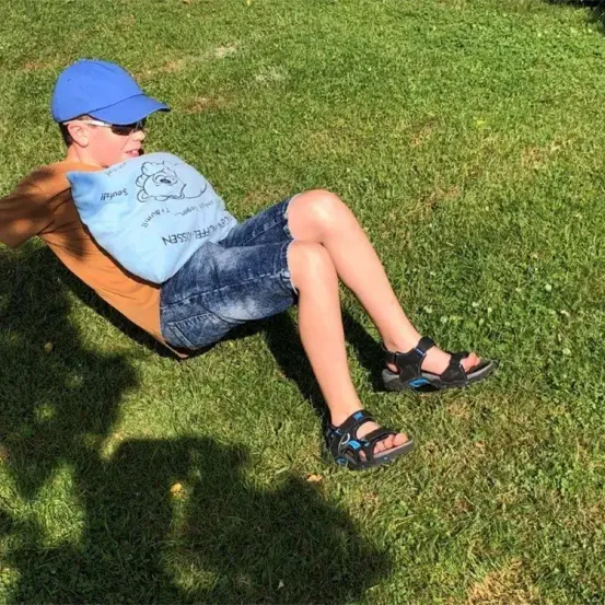 Ein junger Junge, der einen blauen Hut und Sonnenbrille trägt, liegt auf einem Grasfeld und hält ein blaues Kissen.