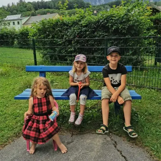 Drei Kinder sitzen auf einer blauen Bank in einem Park. Ein Mädchen hält einen Becher, und ein Junge trägt eine Kappe. Dahinter steht ein grüner Zaun und ein Gebäude.