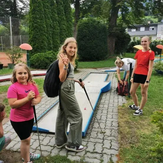 Drei Mädchen spielen Minigolf in einem Park. Das Mädchen in der Mitte lächelt und hält einen Golfschläger. Ein anderes Mädchen beugt sich nach unten, um einen Golfball aufzuheben. Ein anderes Mädchen steht links. Hinter ihnen sind Bäume und ein Haus.