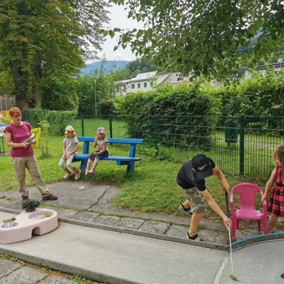Eine Frau steht in der Nähe eines Golfplatzes mit Kindern und einem Jungen, der einen Schläger hält. Dahinter befindet sich eine Bank und ein Mädchen. Im Hintergrund befindet sich ein Zaun und ein Gebäude.
