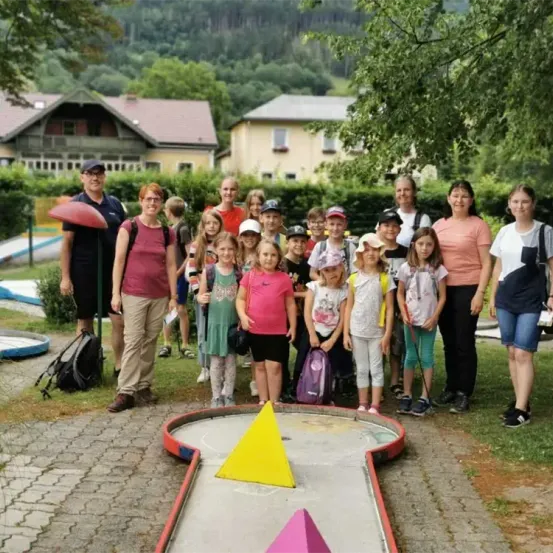Eine Gruppe von Menschen, darunter Kinder, posiert für ein Foto in einem Park. Einige tragen Hüte, Brillen und Turnschuhe. Sie stehen vor einem kleinen Golfplatz. Hinter ihnen sind Häuser und Bäume.