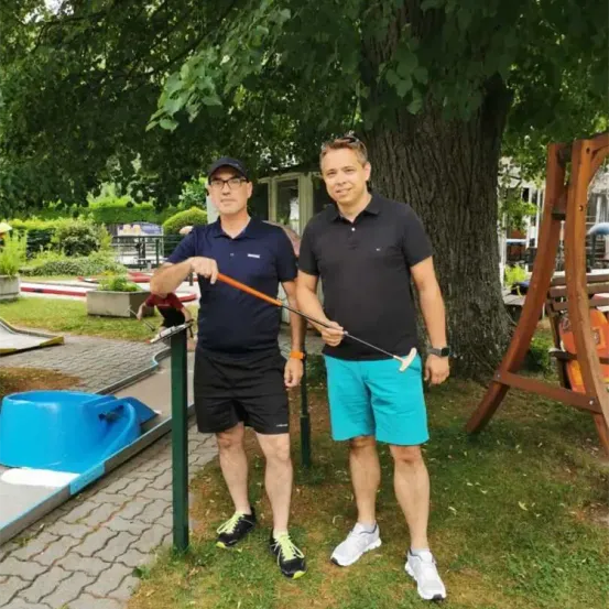 Zwei Männer in Golfkleidung posieren für ein Foto in einem Park. Einer hält einen Golfschläger, der andere einen Stock. Ein Baum und ein Golfplatz sind hinter ihnen.