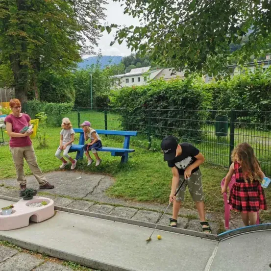 Eine Familie spielt Minigolf. Ein Erwachsener liest ein Buch, ein Junge schlägt einen Ball, und Mädchen sitzen auf einer Bank und schauen zu.