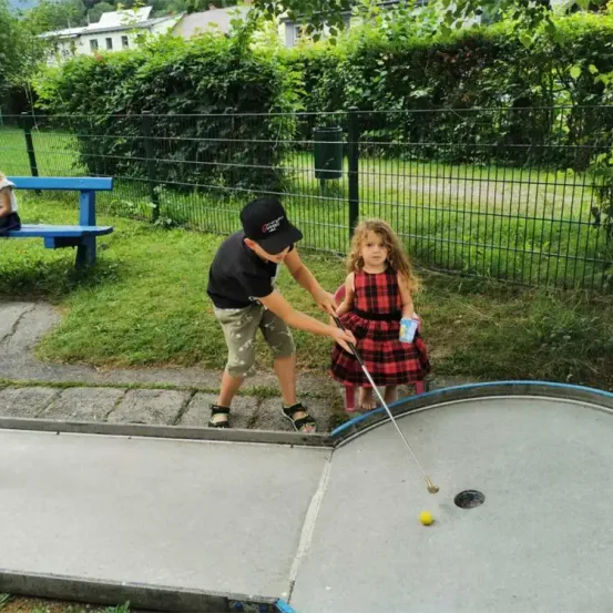 Ein Junge und ein Mädchen spielen Minigolf. Der Junge schlägt den Ball, während das Mädchen zuschaut. Dahinter steht eine Bank und ein Zaun.