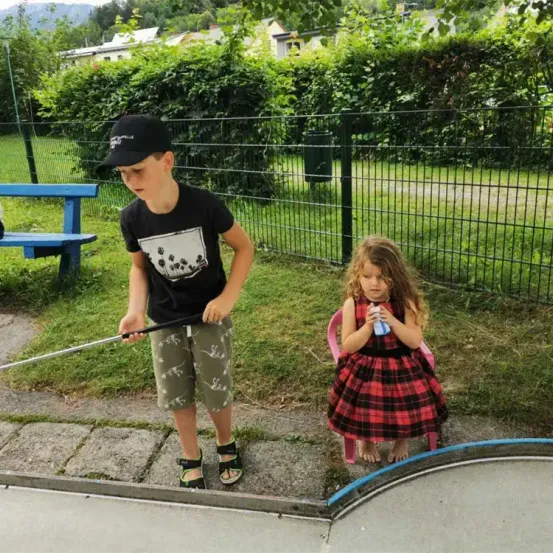 Ein Junge und ein Mädchen sind draußen, der Junge hält einen Golfschläger und das Mädchen sitzt auf einem Stuhl und hält eine Flasche.