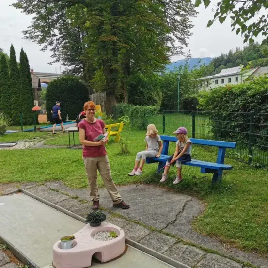 Eine Frau mit einem Buch steht in einem Park mit zwei Kindern auf einer Bank. Dahinter ist ein Golfplatz zu sehen, auf dem eine Person geht. Der Park ist von Grünflächen und einem Zaun umgeben.