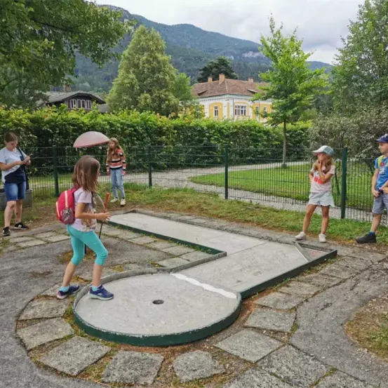 Mehrere Kinder spielen Golf auf einem kleinen Platz im Freien, in der Nähe einiger Erwachsener. Ein Kind mit einem rosa Rucksack hält einen Schläger. Dahinter befindet sich ein eingezäunter Bereich mit Häusern und Bergen.