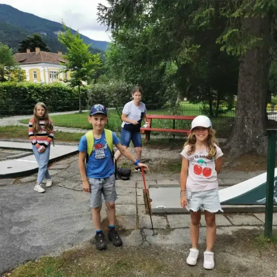 Drei Kinder und ein Erwachsener stehen in einem Park mit einem Golfplatz. Die Kinder lächeln und schauen zur Kamera. Ein Mädchen trägt einen weißen Hut. Hinter ihnen steht eine rote Bank und ein Baum. In der Ferne befindet sich ein Haus mit Bergblick.