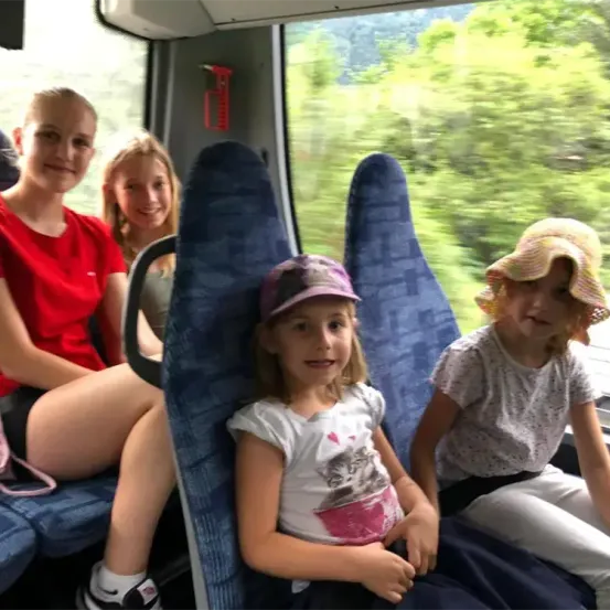 Vier Mädchen sitzen lächelnd auf einem Bus und schauen aus dem Fenster, wobei Bäume draußen sichtbar sind.