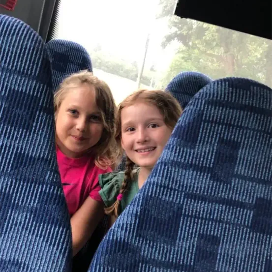 Zwei Mädchen sitzen lächelnd in einem blauen Bus. Eine trägt ein rosa T-Shirt, die andere ein grünes Kleid. Sie schauen in die Kamera, wobei Bäume durch das Busfenster sichtbar sind.