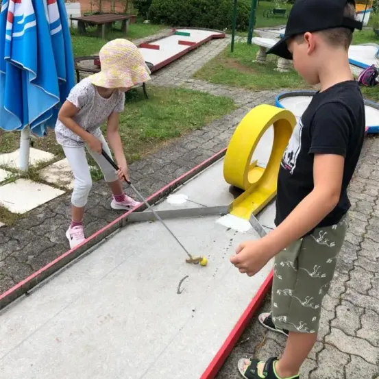 Zwei Kinder spielen Minigolf. Ein Mädchen mit einem gelben Hut und ein Junge in einem schwarzen T-Shirt. Sie schlagen einen gelben Ball auf einem Golfplatz.