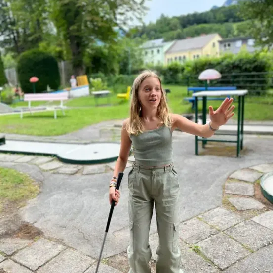 Ein junges Mädchen mit blonden Haaren steht auf einem Betonweg und hält einen Golfschläger in der Hand. Sie lächelt und scheint für ein Foto zu posieren. Hinter ihr befindet sich ein Golfplatz mit einem Mini-Golfplatz, Bänken und einem Tisch. Es gibt auch Bäume und Häuser in der Ferne.