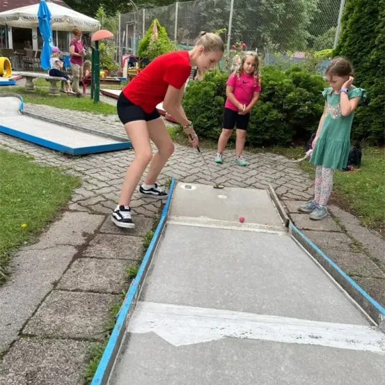 Ein Mädchen in einem roten T-Shirt übt Golf auf einem Grün, während zwei junge Mädchen zuschauen. Dahinter ist ein Spielplatz zu sehen, und ein Gebäude mit einem Schirm im Hintergrund.