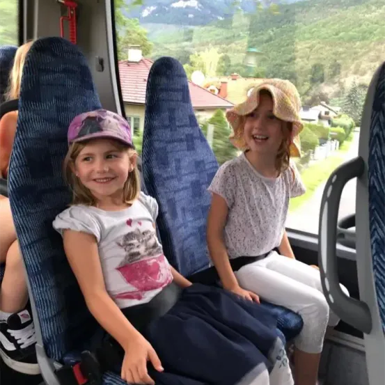 Zwei junge Mädchen sitzen lächelnd in einem Bus, wobei eine eine Mütze und die andere einen Hut trägt. Der Bus ist von blauen Sitzen umgeben und bietet einen malerischen Blick auf ein Haus mit einem roten Dach und einer Bergkulisse.