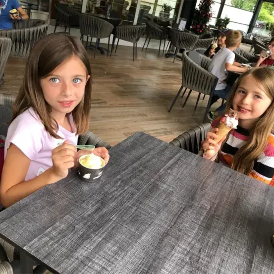 Zwei junge Mädchen sitzen an einem Tisch und essen Eis. Eine hält einen Eisbecher, die andere benutzt einen Löffel. Die Umgebung ist ein Restaurant mit Holzböden und vielen Tischen und Stühlen.