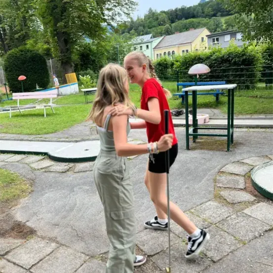 Zwei junge Mädchen umarmen sich in einem Park. Eine hält einen Golfschläger und lächelt, während die andere ein Armband und eine Halskette trägt. Hinter ihnen befindet sich ein Spielplatz mit einer Bank, einem Tisch und einer Lampe.