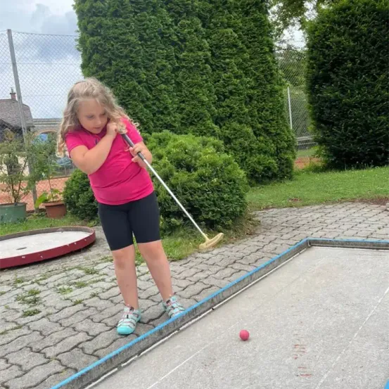 Ein kleines Mädchen in einem rosa Shirt spielt Minigolf auf einem Betonplatz, mit Büschen und einem Zaun im Hintergrund.