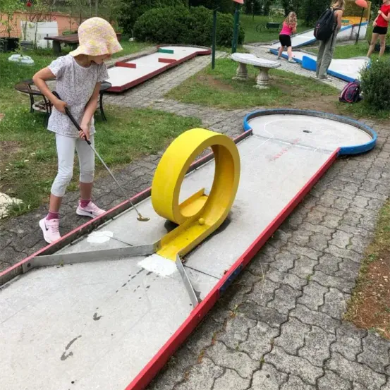 Ein junges Mädchen spielt Minigolf in einem Park und schlägt einen Ball mit einem gelben Schläger in Richtung Loch. Der Platz ist von Grünflächen und anderen Spielern umgeben.