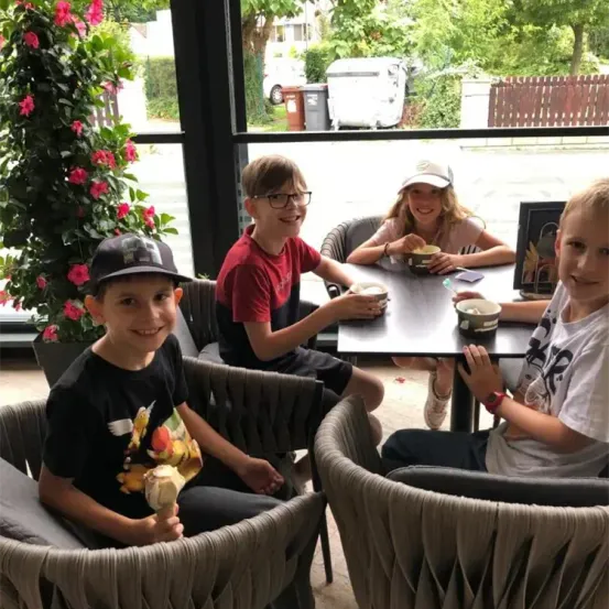 Vier Kinder sitzen um einen Tisch in einem Café und lächeln. Drei halten Eisbecher, eines eine Tasse. Draußen sind ein Baum, ein Mülleimer und ein Auto zu sehen.