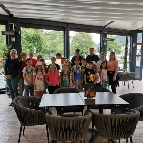 Eine Gruppe von Menschen, darunter Erwachsene und Kinder, posiert für ein Foto in einem Restaurant mit Glaswänden und draußen sichtbaren Bäumen.